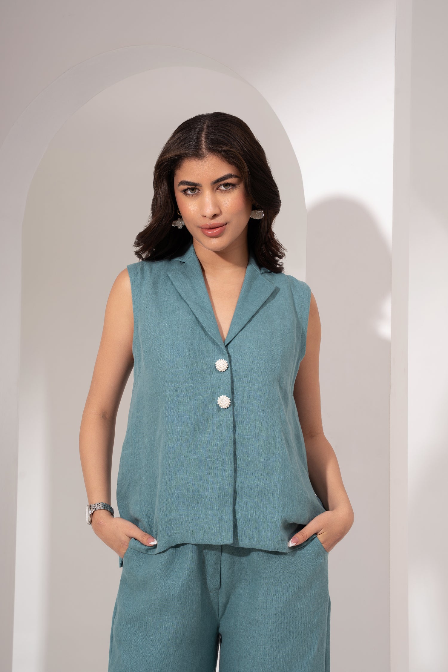 Woman wearing a teal sleeveless blazer and pants against a plain background